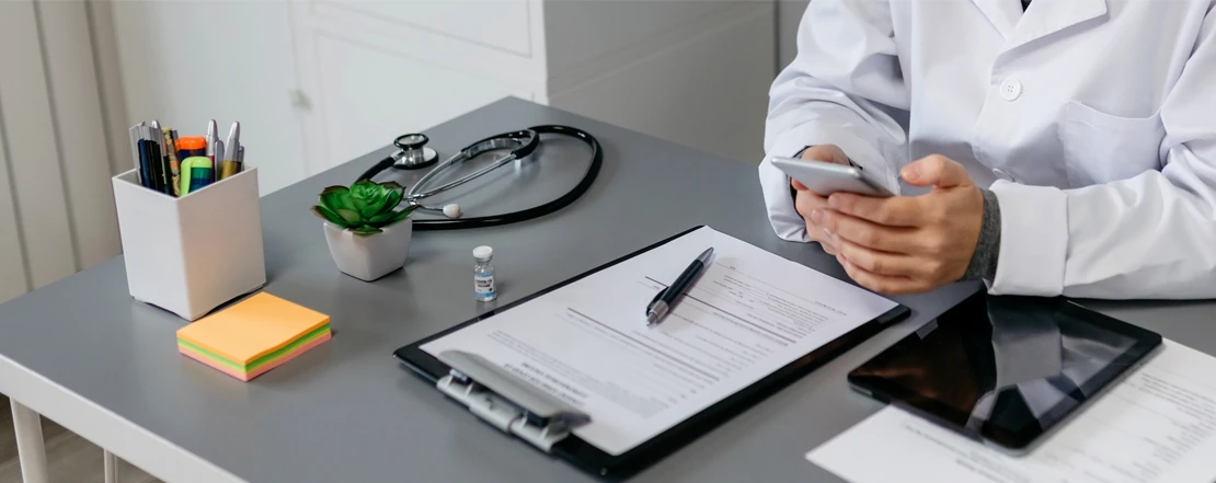 A doctor using a phone at a consultation desk with a stethoscope and medical paperwork
